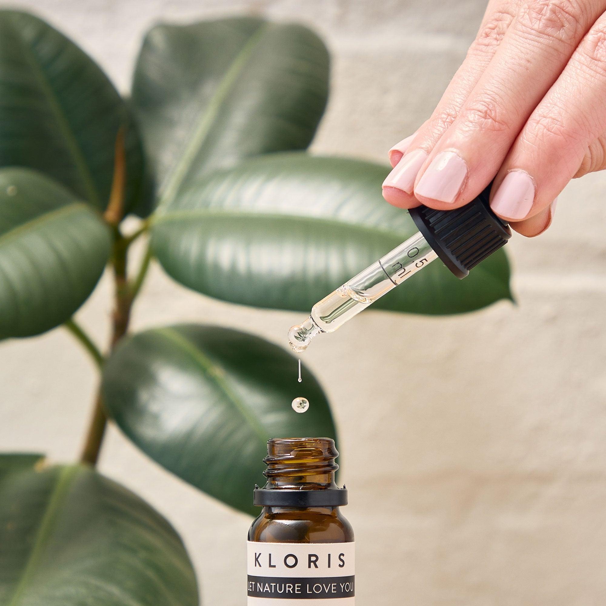 Hand using a dropper to extract a drop from a Kloris bottle with green leaves in the background