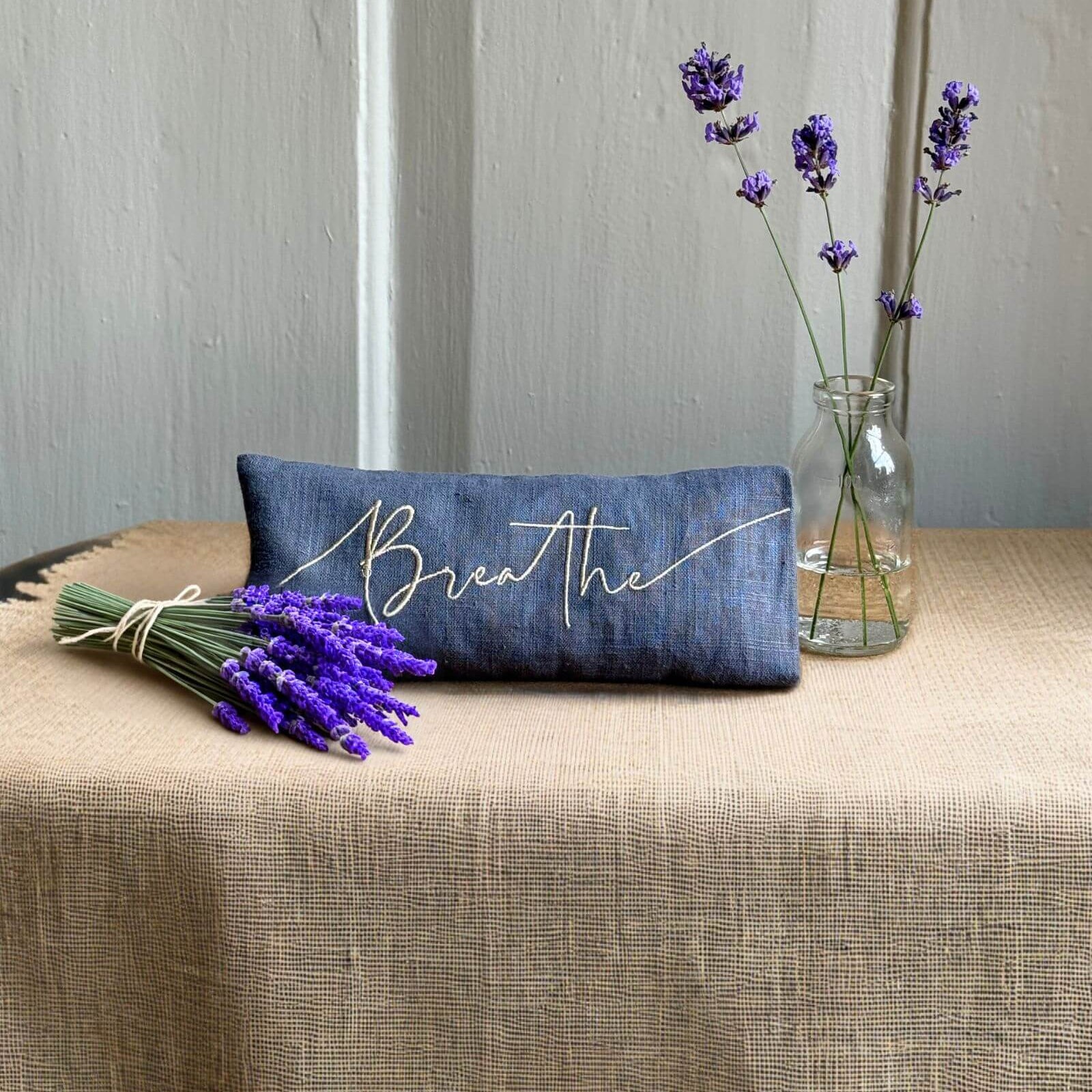 Blue pillow with 'Breathe' text on a table with lavender and a vase against a neutral wall.