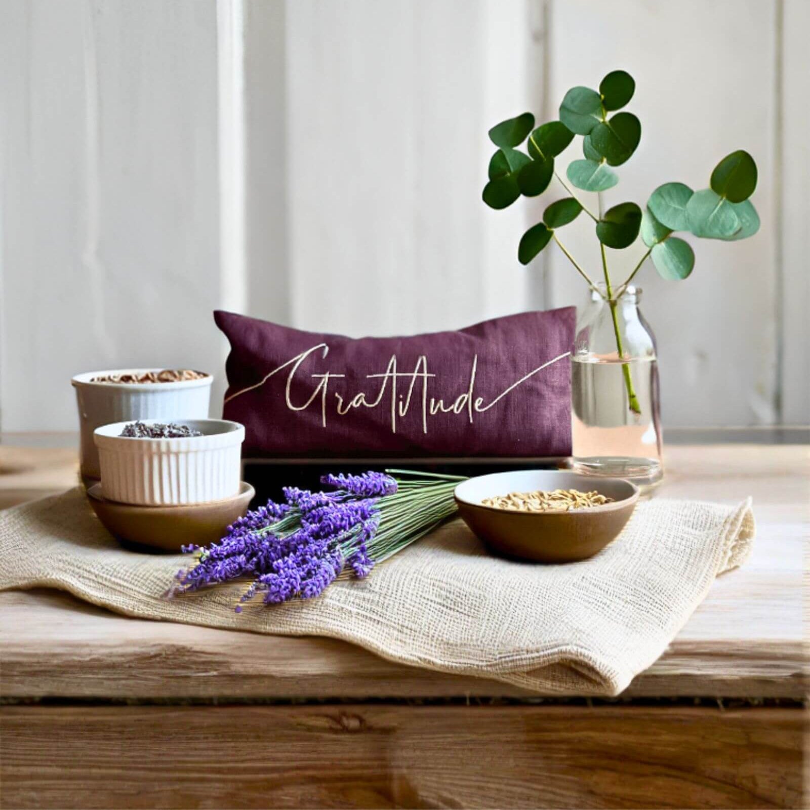 Table setting with a 'Gratitude' pillow, bowls, lavender, and a plant on a wooden surface.