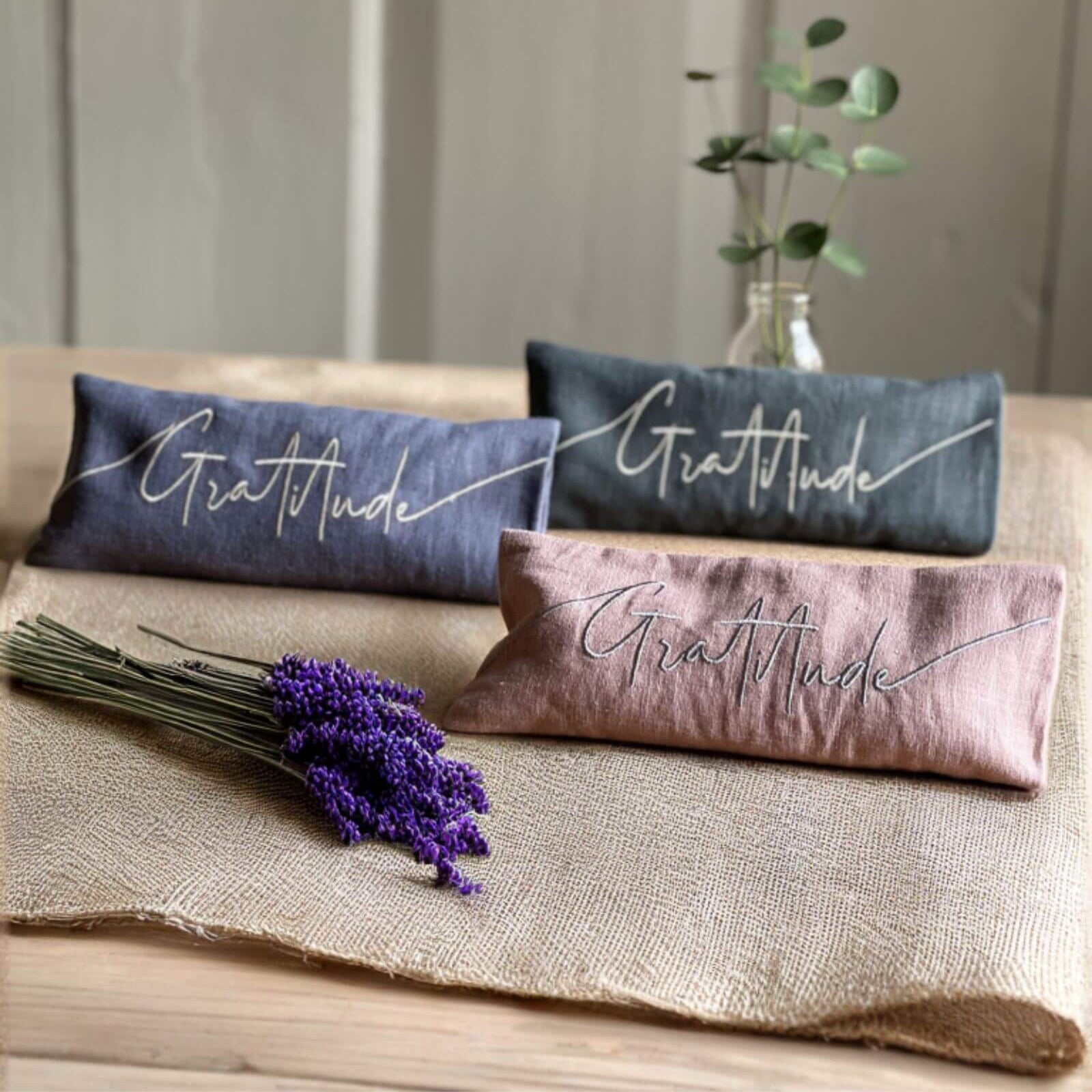 Three 'Gratitude' pillows in different colors on a wooden surface with a plant in the background.