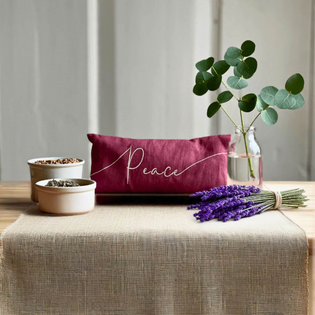Maroon pillow with 'Peace' embroidery on a table with lavender and plants.