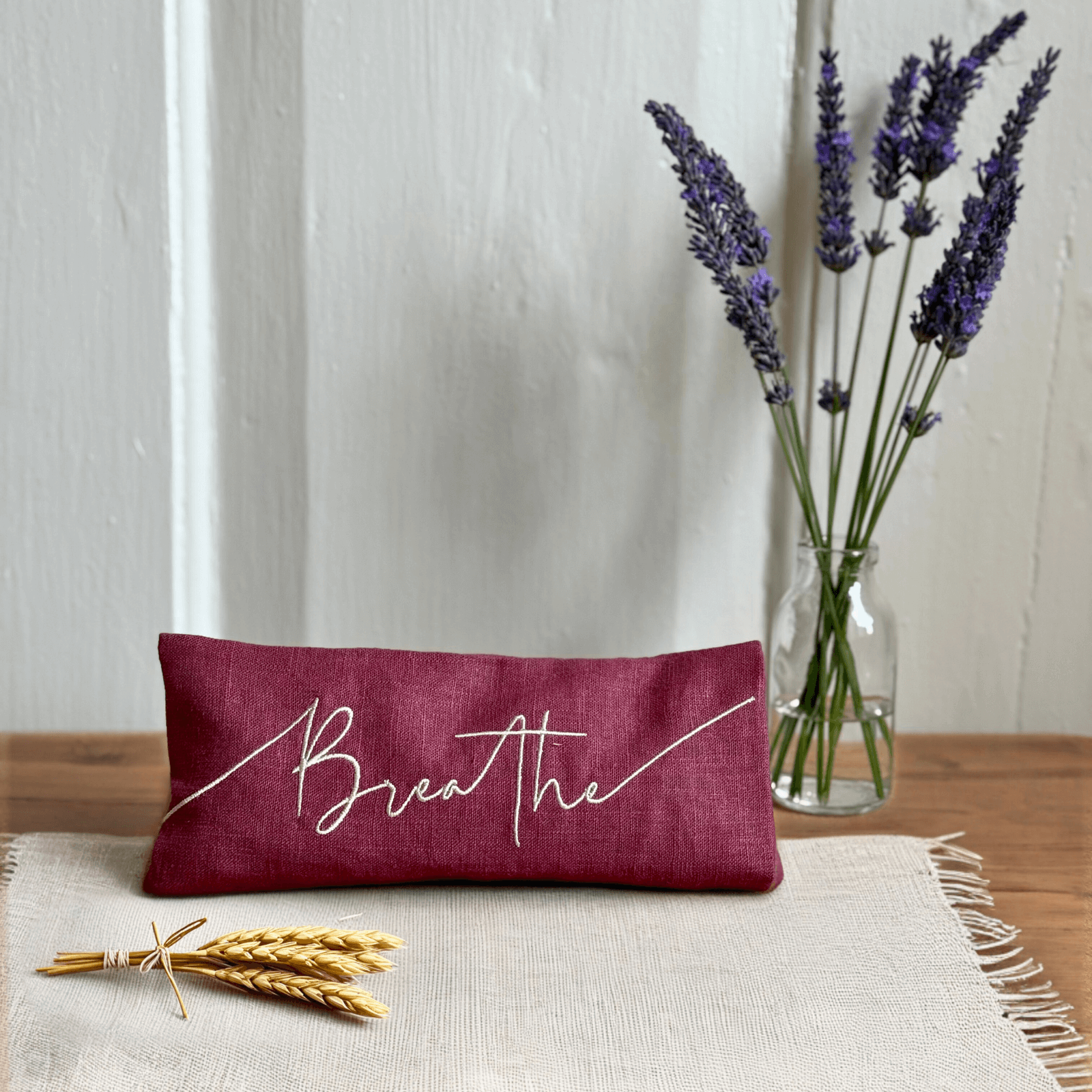 Maroon pillow with 'Breathe' text on a wooden surface with lavender and wheat.