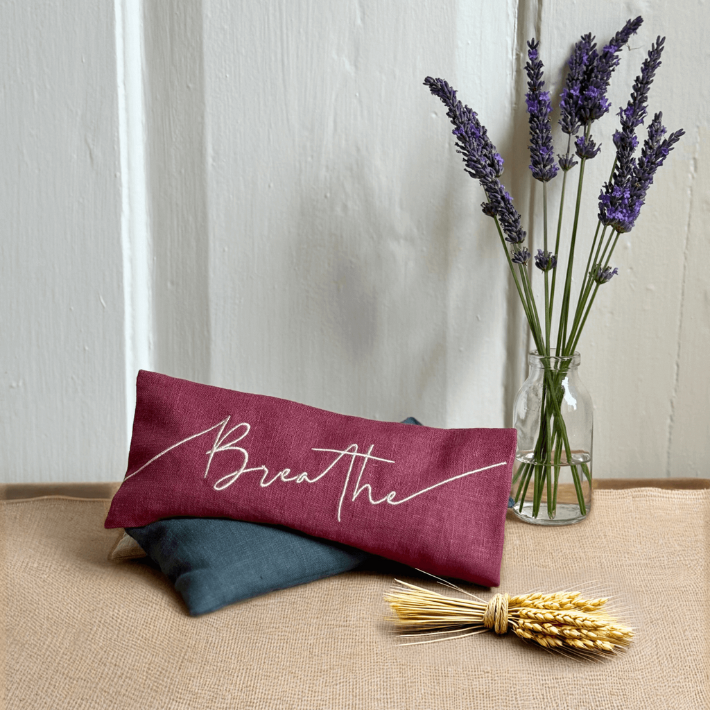 Maroon pillow with 'Breathe' text, glass vase with lavender, and bundle of wheat on a white background
