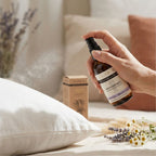 Hand holding a bottle of 'Natura Love' sooting mist over pillows with lavender and flowers.