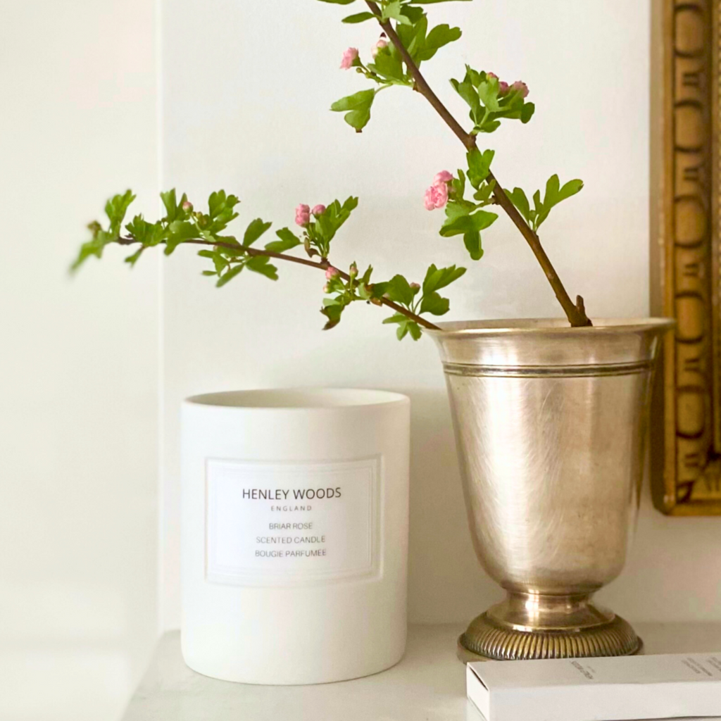 Candle labeled 'Henley Woods' with a decorative plant in a silver vase on a white surface.