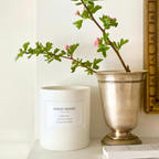Candle labeled 'Henley Woods' with a decorative plant in a silver vase on a white surface.